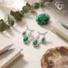 Close-up of pear-cut emerald drop earrings with diamond halos resting on a linen surface beside a green raw crystal