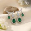 Close-up of pear-cut emerald drop earrings in silver settings with sparkling diamond accents and white floral backdrop