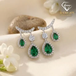 Close-up of pear-cut emerald drop earrings in silver settings with sparkling diamond accents and white floral backdrop