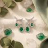 Close-up of pear-cut emerald drop earrings in silver with diamond accents resting on a neutral linen fabric surface