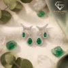 Close-up of pear-cut emerald drop earrings in silver with diamond accents resting on a neutral linen fabric surface