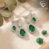 Close-up of pear-cut emerald drop earrings with shimmering diamond halos resting on a soft cream fabric surface