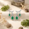 Close-up of pear-cut emerald drop earrings in silver with diamond halos, resting on beige fabric among dried flowers