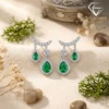 Close-up of pear-cut emerald drop earrings in silver with diamond halos, resting on beige fabric among dried flowers