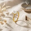Close-up of a gold plated radiant cut engagement ring standing upright on a textured cream fabric with soft sunlight shadows