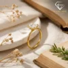 Close-up of a gold plated radiant cut engagement ring resting on a white ceramic plate beside an open vintage book