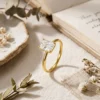 Close-up of a gold plated radiant cut engagement ring resting on linen fabric beside an open vintage book and dried flowers