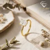 Close-up of a gold plated radiant cut engagement ring resting on linen fabric beside an open vintage book and dried flowers