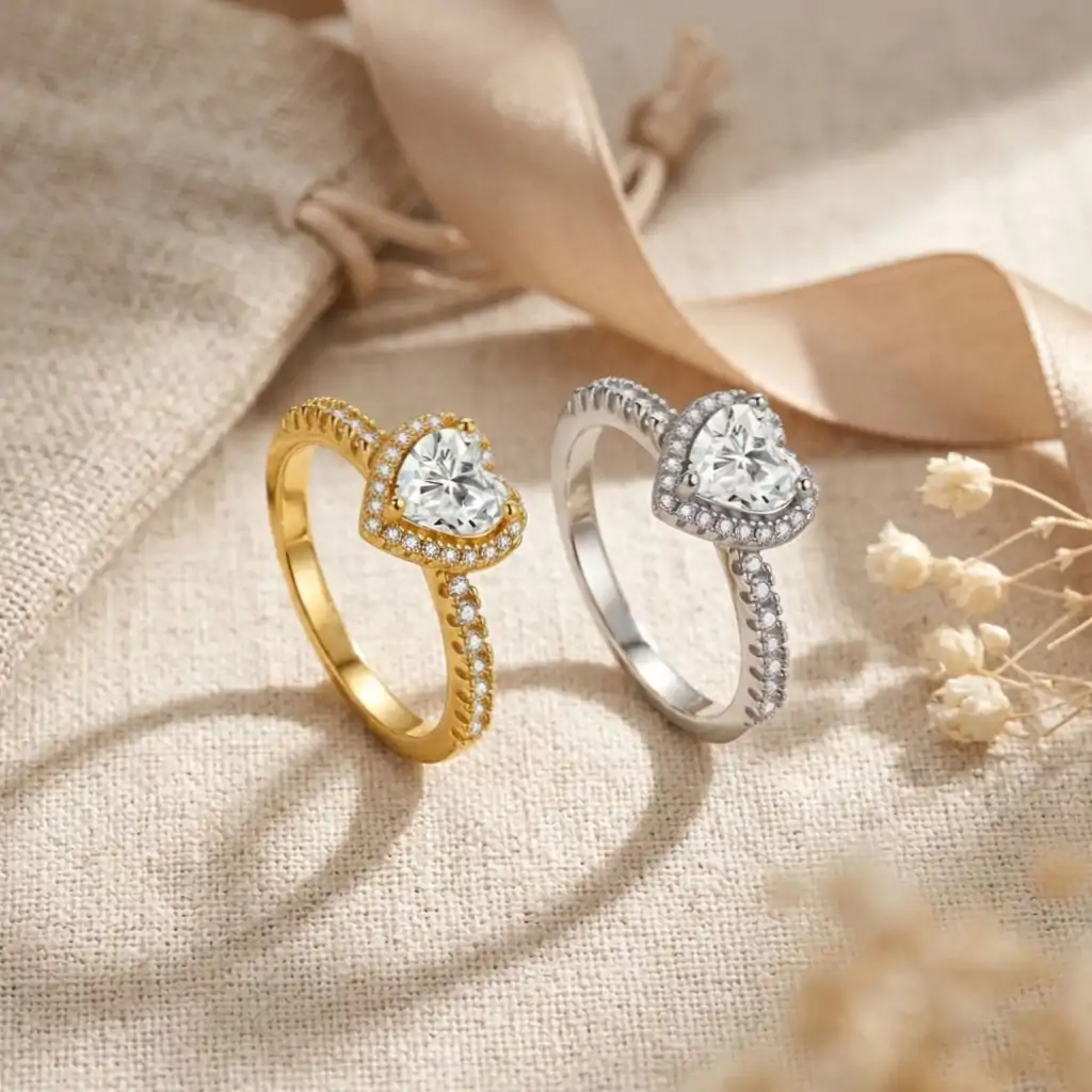 Halo Heart Ring 15 Two moissanite heart ring halo designs side by side, one silver and one gold, both featuring sparkling heart-shaped stones and diamond-encrusted bands, resting on textured beige fabric with soft ribbon and dried flowers