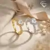 Halo Heart Ring 2 Two moissanite heart ring halo designs side by side, one silver and one gold, both featuring sparkling heart-shaped stones and diamond-encrusted bands, resting on textured beige fabric with soft ribbon and dried flowers