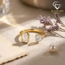Close-up of a gold moissanite toi moi ring featuring one round and one emerald cut stone set on a textured beige surface