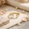 Close-up of a gold moissanite toi moi ring featuring a round and emerald cut stone on a beige fabric with pearls