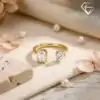 Close-up of a gold moissanite toi moi ring featuring a round and emerald cut stone on a beige fabric with pearls